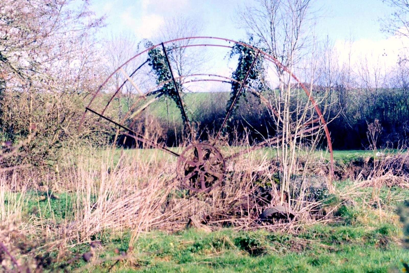 The surviving iron rim of a 1930s waterwheel standing in a field, overgrown with ivy and winter grass